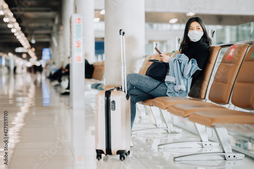 Asian travler woman wearing face mask sitting on social distancing chiar with luggage waiting for flight at airport terminal. tourism reopen after covid pandemic photo