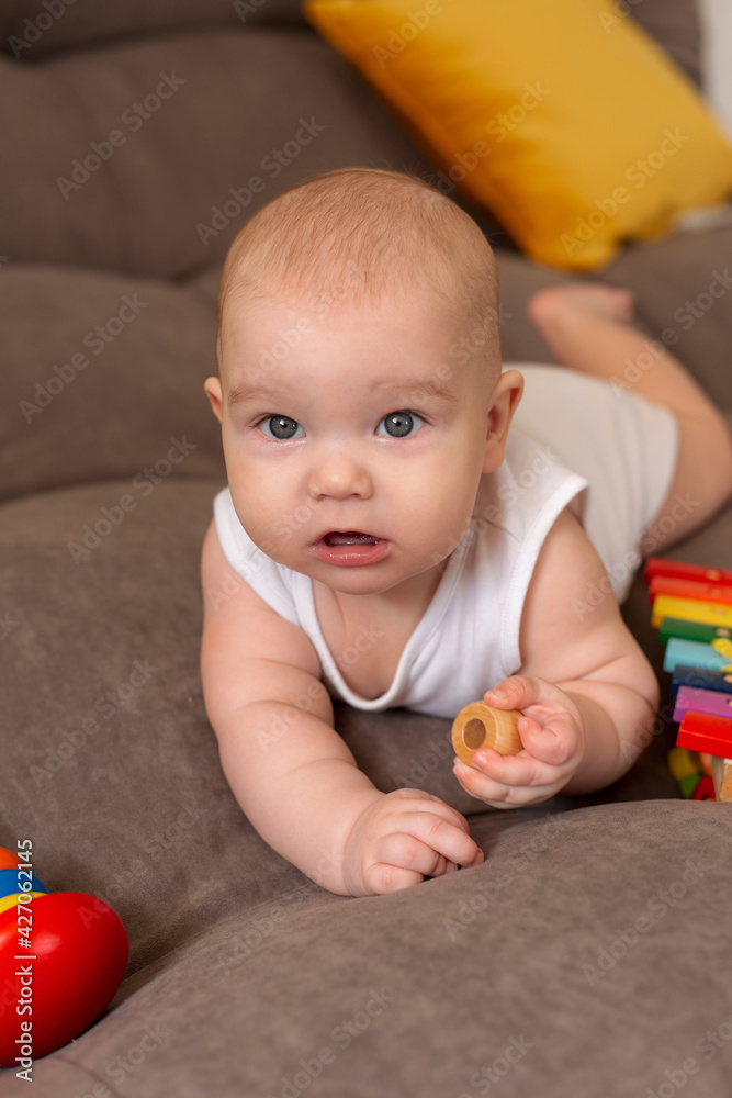 Cute toddler in white bodysuit lies at home on grey sofa with yellow pillows playing with wooden developing toy Cute toddler in white bodysuit lies at home on grey sofa with yellow pillows playing with wooden developing toy
