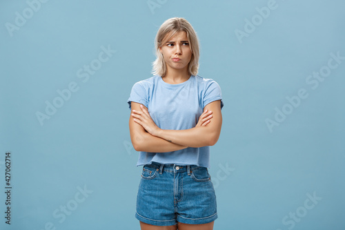 Uncertain troubled and perplexed attractive blond woman with tanned skin holding hands crossed on chest pouting and frowning looking right with worried unsure and doubtful look over blue wall photo