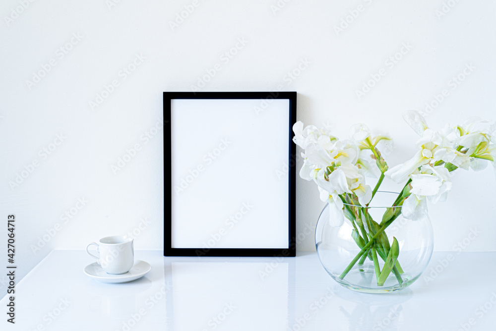 Blank black frame mockup. Fresh flowers of white irises in glass vase in the shape of a sphere on a white table and coffee cup. White background, minimal and elegant space