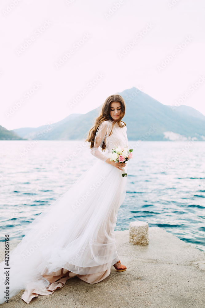 The bride holds a bouquet of roses in her hands and stands on the pier in the Bay of Kotor