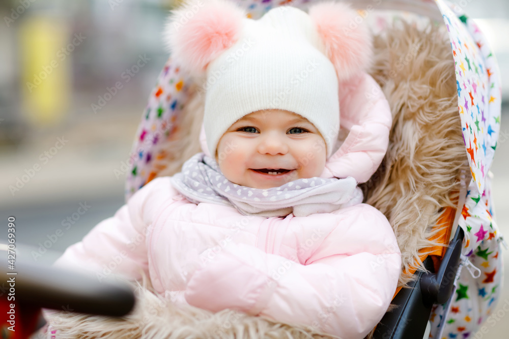 Cute little beautiful baby girl sitting in the pram or stroller on cold autumn, winter or spring day. Happy smiling child in warm clothes, fashion stylish baby coat and hat. Snow falling down Cute little beautiful baby girl sitting in the pram or stroller on cold autumn, winter or spring day. Happy smiling child in warm clothes, fashion stylish baby coat and hat. Snow falling down