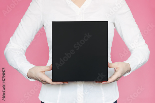 A square black box with an copy empty space for text and design in the hands of a young woman in a white shirt on a pink background. Board, blank, template, mockup, layout for a slogan or inscription