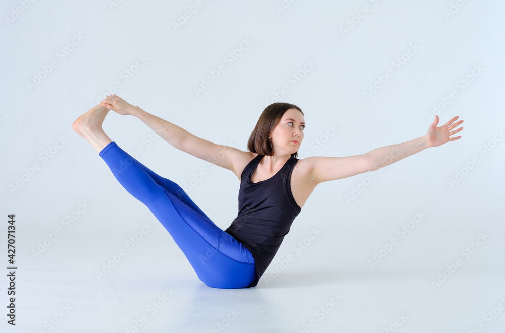 Brunette woman doing yoga exercise Revolved Boat Pose with twist in blue sports leggings and black top, on white background isolated on cyclorama. Parivrtta Navasana.