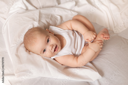Cute toddler in white bodysuit lies in bed with white bedding