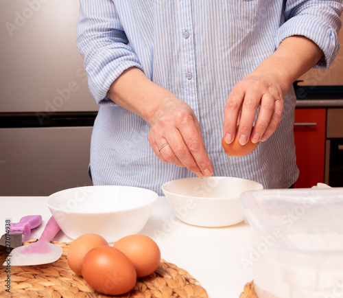 The hands of an aged woman breaks an egg into a white bowl. Selective focus.