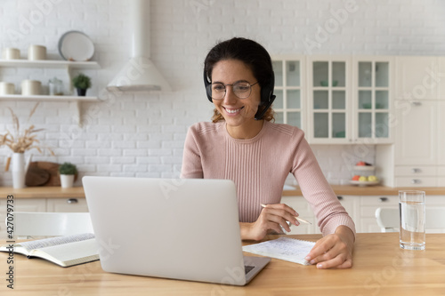 Pleasant elearning. Hispanic female student in earphones study from home look on pc screen listen to tutor teacher prepare to take notes. Smiling young woman enjoy watching webinar online using laptop