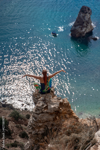The summer lifestyle of a happy stunning woman sitting on a rock above the sea. Enjoying life and looking at the sea. Turquoise sea background. In colored clothes.