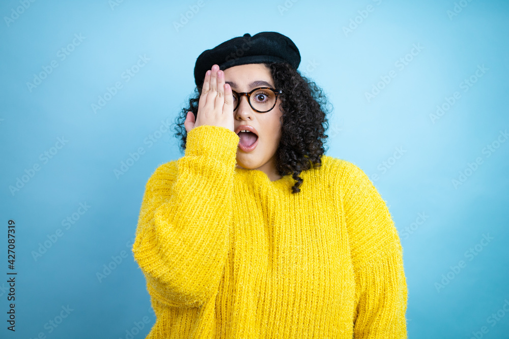 Young beautiful woman wearing french look with beret and yellow casual sweater over isolated blue background covering one eye with hand, confident smile on face and surprise emotion.