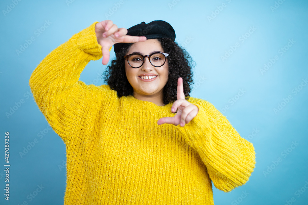Young beautiful woman wearing french look with beret and yellow casual sweater over isolated blue background smiling making frame with hands and fingers with happy face