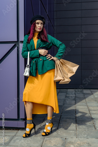 A happy shopaholic girl keeps her bags near the shopping center. A woman near the store is happy with her purchases, holding bags. She is wearing a yellow dress and a green jacket. Consumer concept.