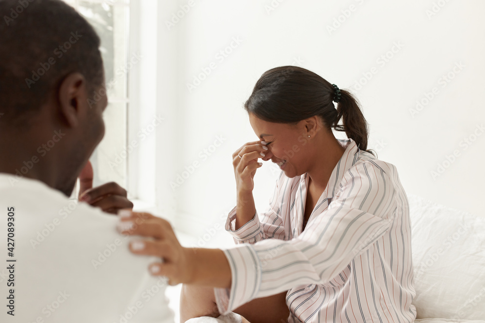 People, domestic life and marriage concept. Portrait of adorable young mixed race female wearing silk pajamas sitting in bedroom, enjoying lazy morning with her husband, laughing, having fun People, domestic life and marriage concept. Portrait of adorable young mixed race female wearing silk pajamas sitting in bedroom, enjoying lazy morning with her husband, laughing, having fun