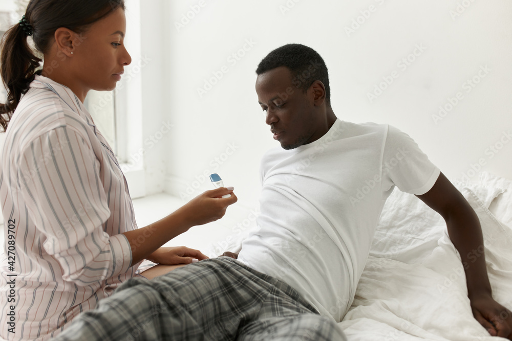 Obraz premium Portrait of frustrated young African man lying on bed his unhappy girlfriend showing him pregnancy test stick with positive result, saying I'm pregnant, worrying about future consequences