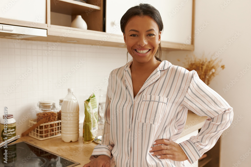 Home, domesticity and cooking. Attractive joyful young Hispanic female wearing striped pajamas standing in stylish kitchen interior, smiling confidently, making coffee, having breakfast indoors Home, domesticity and cooking. Attractive joyful young Hispanic female wearing striped pajamas standing in stylish kitchen interior, smiling confidently, making coffee, having breakfast indoors