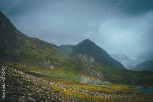 Dramatic rainy alpine landscape with low rainbow in green valley and dark sharp pinnacle in low clouds. Pointed rocks in overcast weather. Atmospheric awesome view to pointy mountain in low clouds. © Daniil