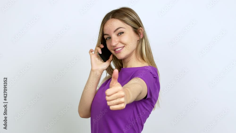 Young woman talking on mobile phone giving thumbs up and smiling because something good has happened over isolated background Young woman talking on mobile phone giving thumbs up and smiling because something good has happened over isolated background