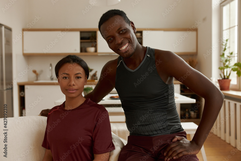 Obraz premium Cheerful handsome young Afro American male sitting on comfortable couch next to his attractive wife wearing sports clothing, going out for running exercise. People, relationships and lifestyle concept