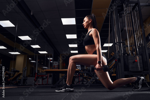 Slim fit woman doing lunges with dumbbells in her hands in a modern gym