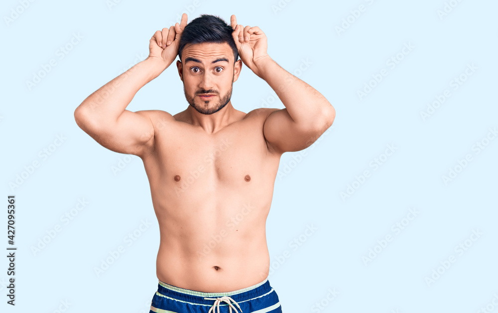 Young handsome man wearing swimwear doing funny gesture with finger over head as bull horns