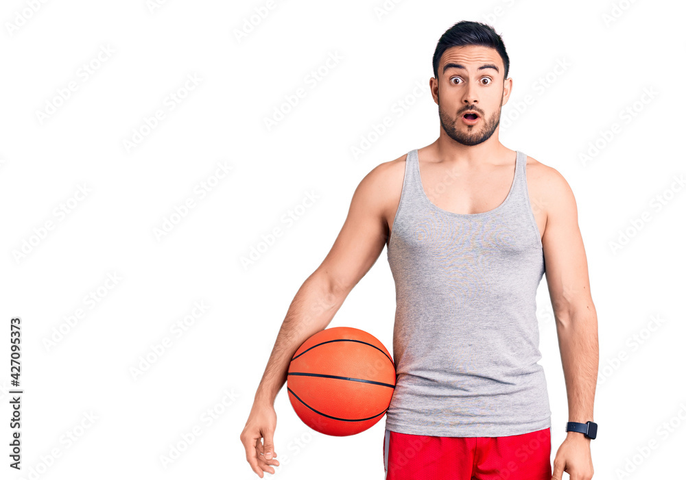 Young handsome man holding basketball ball scared and amazed with open mouth for surprise, disbelief face Young handsome man holding basketball ball scared and amazed with open mouth for surprise, disbelief face