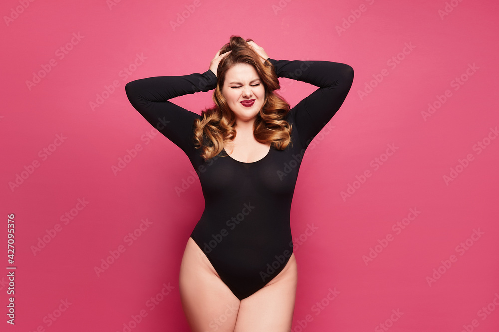 Obraz premium Beautiful emotional plus-size female model in a black bodysuit posing on pink background in studio