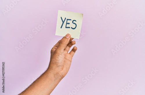 Hand of hispanic man holding yes reminder paper over isolated pink background.