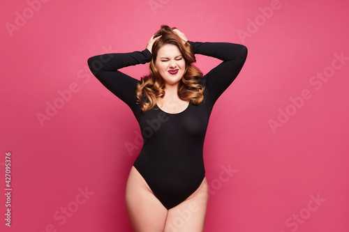 Beautiful emotional plus-size female model in a black bodysuit posing on pink background in studio © innarevyako
