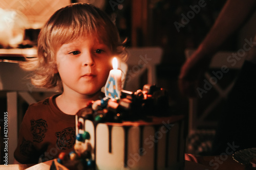 A cute four year old kid blows out the candle on his birthday cake  which is on the table  the lights are off. Children s party. A little boy s birthday celebration. Family having fun at happy holiday