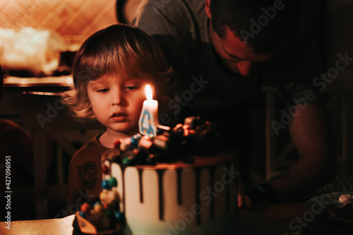 A cute four year old kid blows out the candle on his birthday cake, which is on the table, the lights are off. Children's party. A little boy's birthday celebration. Family having fun at happy holiday
