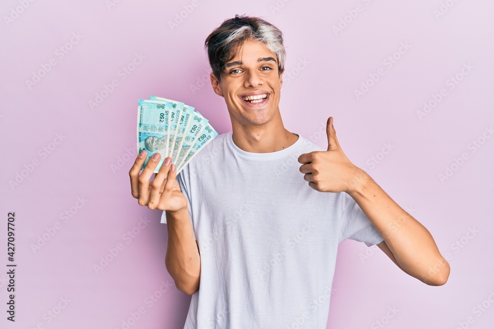 Young hispanic man holding 50 indian rupee banknotes smiling happy and positive, thumb up doing excellent and approval sign Young hispanic man holding 50 indian rupee banknotes smiling happy and positive, thumb up doing excellent and approval sign