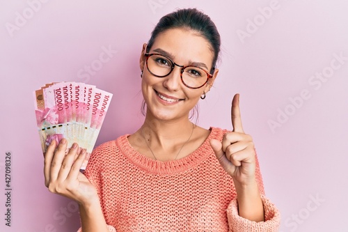 Young caucasian woman holding 100000 indonesian rupiah smiling with an idea or question pointing finger with happy face, number one photo