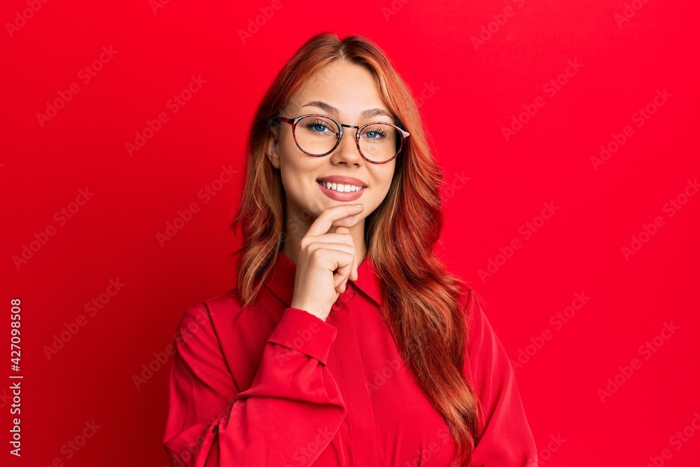 Young beautiful redhead woman wearing casual clothes and glasses over red background looking confident at the camera with smile with crossed arms and hand raised on chin. thinking positive. Young beautiful redhead woman wearing casual clothes and glasses over red background looking confident at the camera with smile with crossed arms and hand raised on chin. thinking positive.