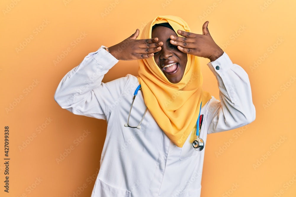 Beautiful african young woman wearing doctor uniform and hijab covering eyes with hands smiling cheerful and funny. blind concept.