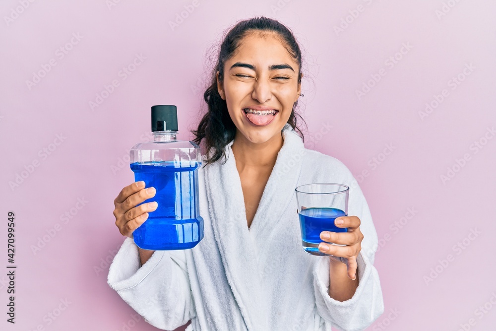 Hispanic teenager girl with dental braces holding mouthwash sticking tongue out happy with funny expression. Hispanic teenager girl with dental braces holding mouthwash sticking tongue out happy with funny expression.