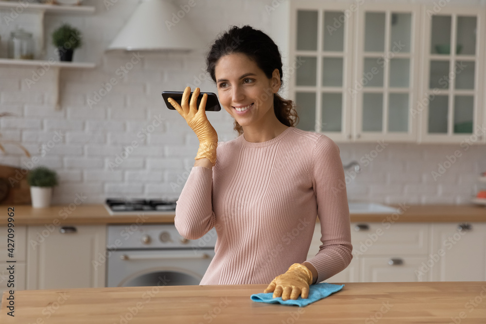 Modern day housewife. Young latina female engaged in cleaning modern kitchen remove wipe dust wear rubber gloves to protect hands hold mobile phone use voice dialing listen to caller by loudspeaker Modern day housewife. Young latina female engaged in cleaning modern kitchen remove wipe dust wear rubber gloves to protect hands hold mobile phone use voice dialing listen to caller by loudspeaker