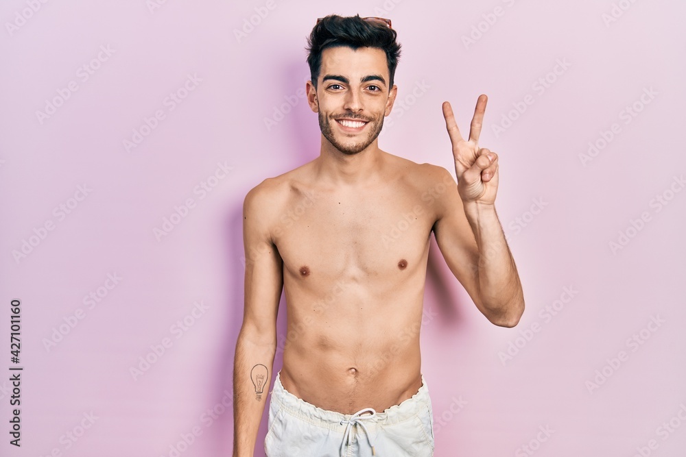 Young hispanic man wearing swimwear shirtless showing and pointing up with fingers number two while smiling confident and happy.