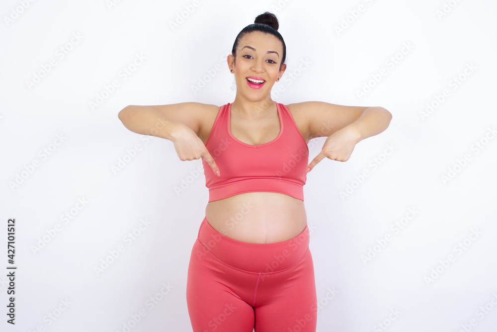 young beautiful Arab pregnant woman in sports clothes against white wall with positive expression, points down with both index fingers, has broad interested smile. Look there, please. young beautiful Arab pregnant woman in sports clothes against white wall with positive expression, points down with both index fingers, has broad interested smile. Look there, please.