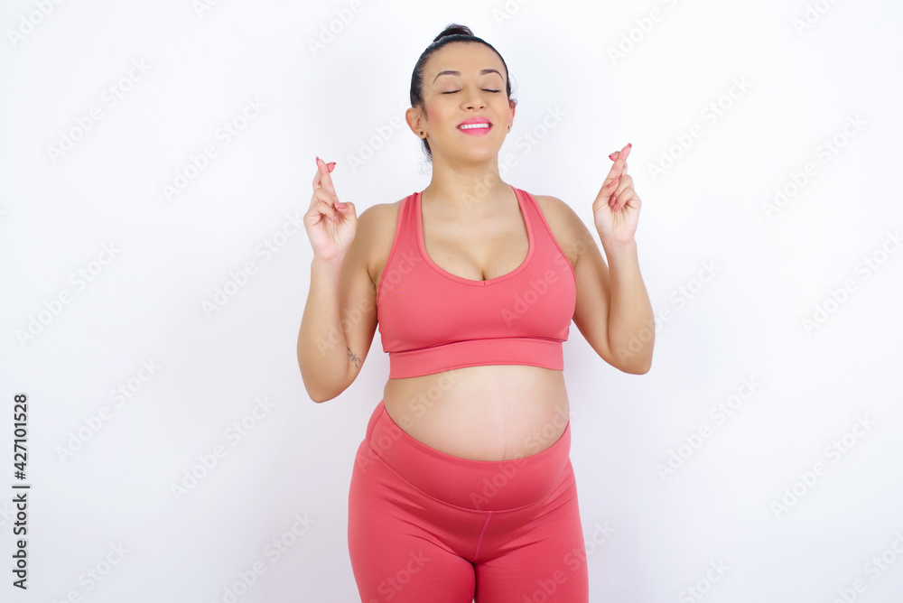 Joyful young beautiful Arab pregnant woman in sports clothes against white wall clenches teeth, raises fingers crossed, makes desirable wish, waits for good news, I have to win. Joyful young beautiful Arab pregnant woman in sports clothes against white wall clenches teeth, raises fingers crossed, makes desirable wish, waits for good news, I have to win.