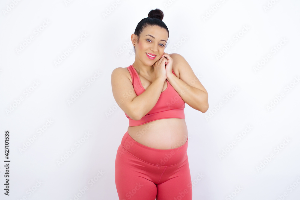 Dreamy young beautiful Arab pregnant woman in sports clothes against white wall with pleasant expression, closes eyes, keeps hands crossed near face, thinks about something pleasant Dreamy young beautiful Arab pregnant woman in sports clothes against white wall with pleasant expression, closes eyes, keeps hands crossed near face, thinks about something pleasant