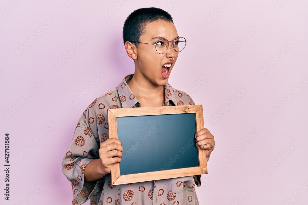 Beautiful hispanic woman with short hair holding blackboard angry and mad screaming frustrated and furious, shouting with anger. rage and aggressive concept. Beautiful hispanic woman with short hair holding blackboard angry and mad screaming frustrated and furious, shouting with anger. rage and aggressive concept.