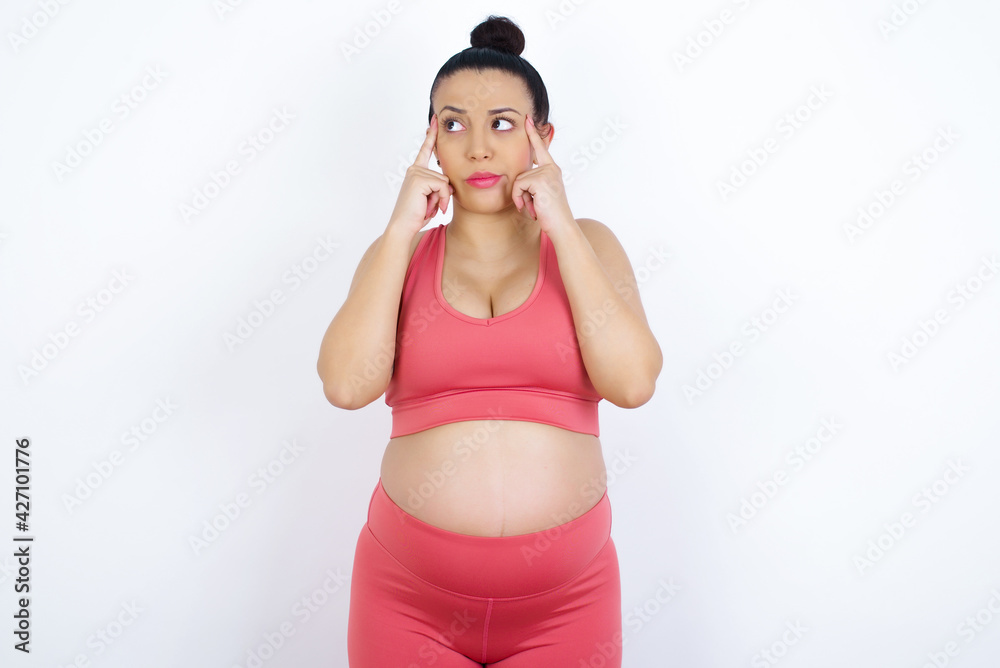 young beautiful Arab pregnant woman in sports clothes against white wall with thoughtful expression, looks away, keeps hand near face, thinks about something pleasant. young beautiful Arab pregnant woman in sports clothes against white wall with thoughtful expression, looks away, keeps hand near face, thinks about something pleasant.