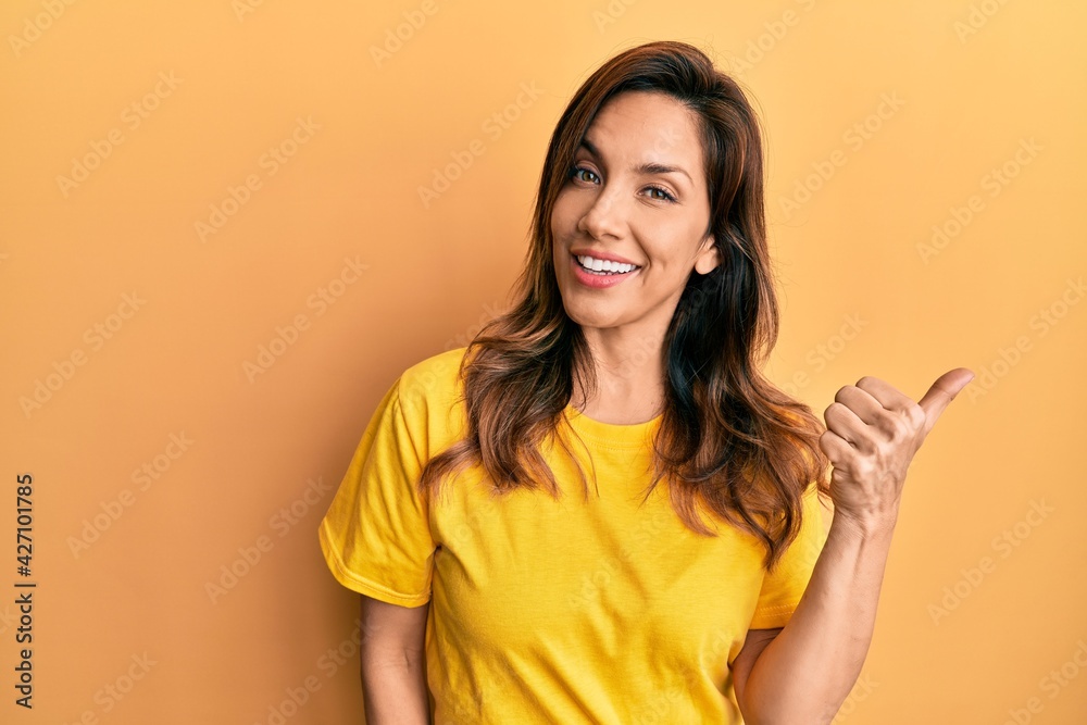 Young latin woman wearing casual clothes smiling with happy face looking and pointing to the side with thumb up. Young latin woman wearing casual clothes smiling with happy face looking and pointing to the side with thumb up.