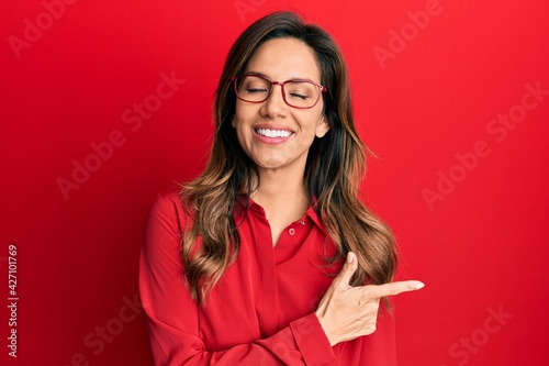 Young latin woman wearing casual clothes and glasses cheerful with a smile of face pointing with hand and finger up to the side with happy and natural expression on face