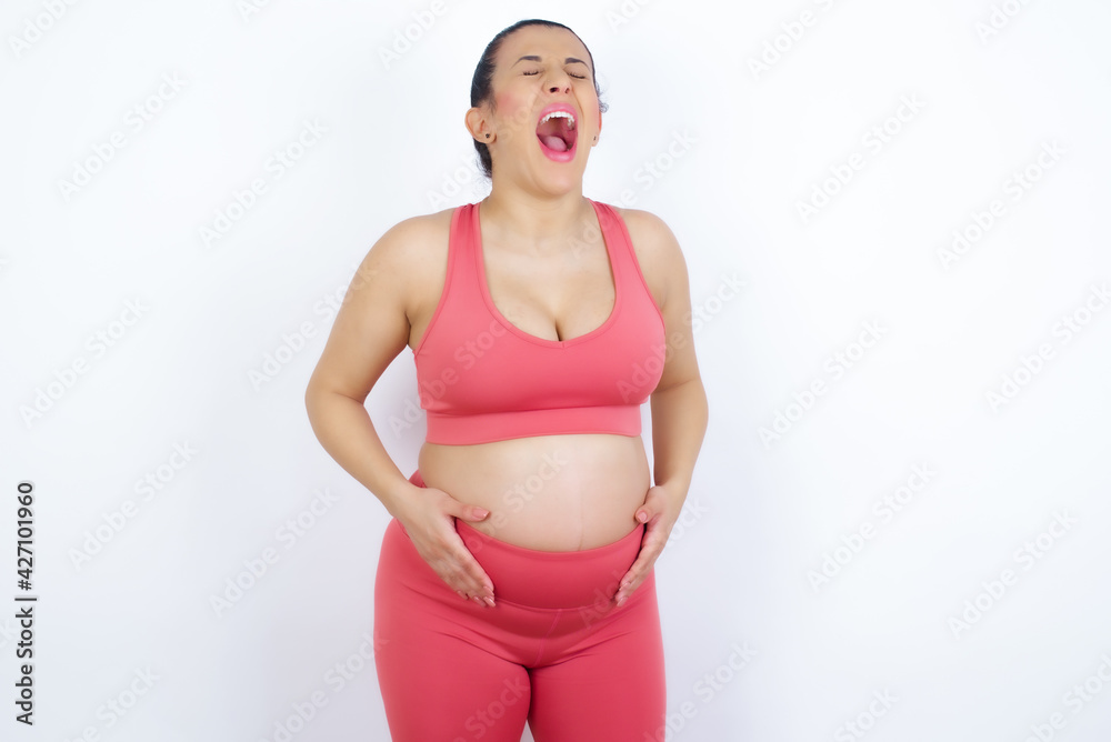 Stressful young beautiful Arab pregnant woman in sports clothes against white wall screams in panic, closes eyes in terror, keeps hands on head, finds out terrified news, can't believe it. Stressful young beautiful Arab pregnant woman in sports clothes against white wall screams in panic, closes eyes in terror, keeps hands on head, finds out terrified news, can't believe it.