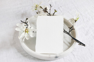 Wedding stationery mock-up scene. Blank vertical greeting card on marble tray. White linen tablecloth background with blooming magnolia stellata tree branches. Feminine still life. Flat lay, top view.
