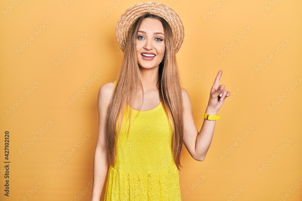 Young blonde girl wearing summer hat with a big smile on face, pointing with hand finger to the side looking at the camera.