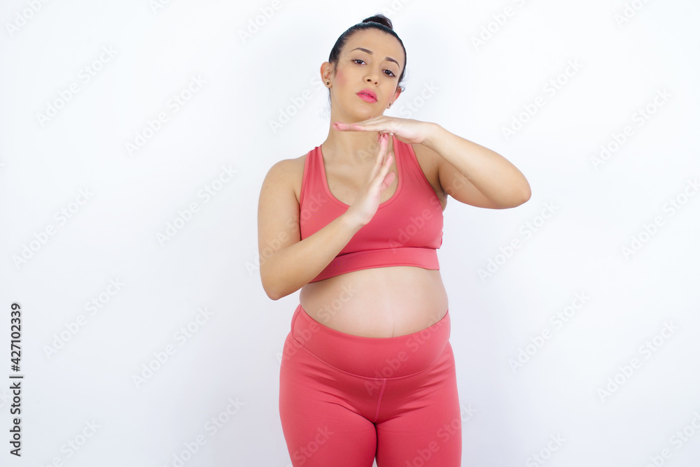 young beautiful Arab pregnant woman in sports clothes against white wall feels tired and bored, making a timeout gesture, needs to stop because of work stress, time concept. young beautiful Arab pregnant woman in sports clothes against white wall feels tired and bored, making a timeout gesture, needs to stop because of work stress, time concept.