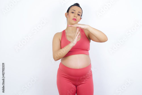 young beautiful Arab pregnant woman in sports clothes against white wall feels tired and bored, making a timeout gesture, needs to stop because of work stress, time concept.