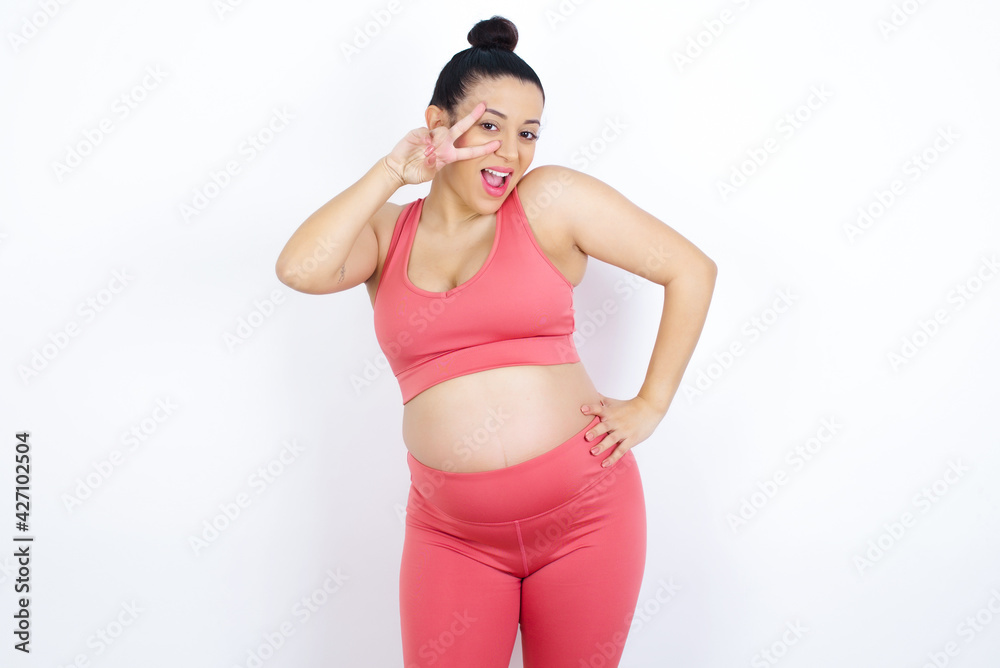young beautiful Arab pregnant woman in sports clothes against white wall making v-sign near eyes. Leisure, coquettish, celebration, and flirt concept. young beautiful Arab pregnant woman in sports clothes against white wall making v-sign near eyes. Leisure, coquettish, celebration, and flirt concept.