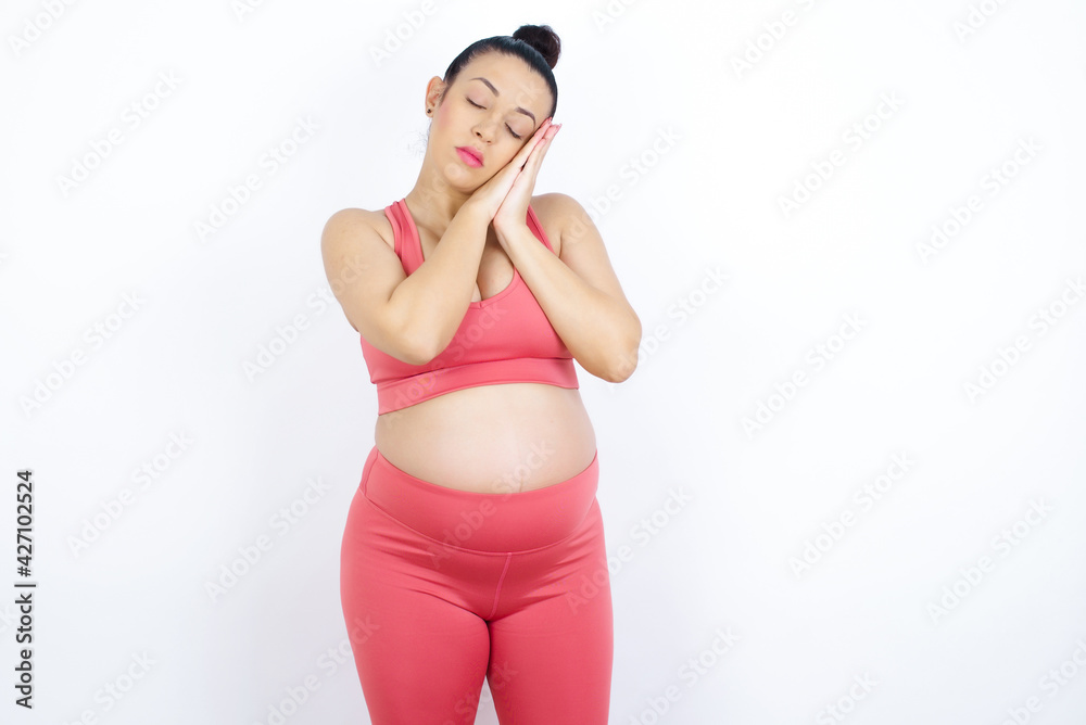 Relax and sleep time. Tired young beautiful Arab pregnant woman in sports clothes against white wall with closed eyes leaning on palms making sleeping gesture. Relax and sleep time. Tired young beautiful Arab pregnant woman in sports clothes against white wall with closed eyes leaning on palms making sleeping gesture.